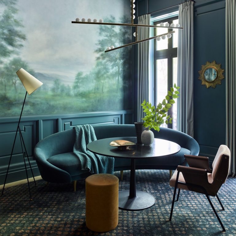 A stylish West Village Townhouse living room with teal walls, a nature mural, blue velvet sofa, round black table, modern brown chair, floor lamp, gold-framed mirror, tall curtains, leafy plant in a vase, and contemporary chandelier with white globe bulbs.