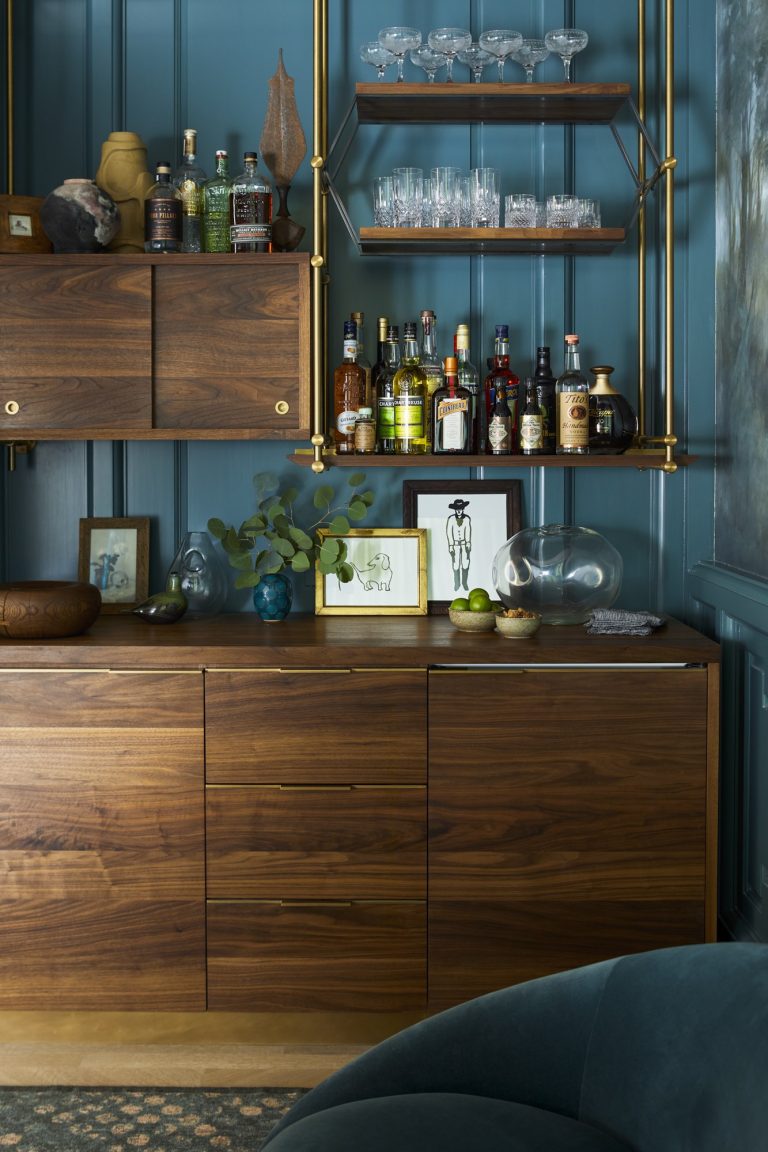 A modern home bar in a West Village Townhouse features teal paneled walls, wooden cabinetry, shelves with assorted liquor bottles, glassware, green foliage in a vase, and gold accents. Moody lighting adds an elegant New York ambiance.