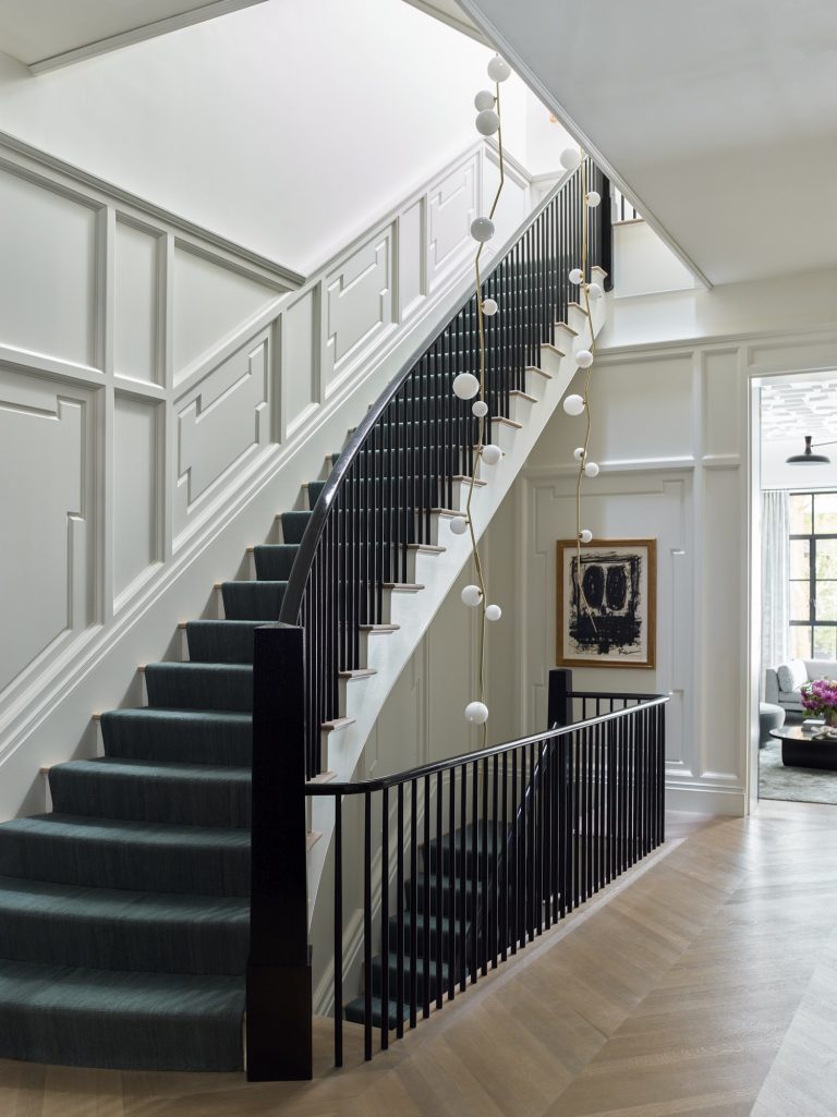 A modern staircase in this West Village Townhouse features dark handrails, green carpeted steps, and white paneled walls. Spherical pendant lights hang alongside the stairs, with abstract artwork and herringbone wood flooring throughout.