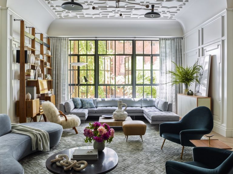 A modern living room in a West Village Townhouse featuring a large gray sectional sofa, blue velvet chairs, a round coffee table with flowers, floor-to-ceiling windows, abstract decor, and bookshelves. Natural light fills the space.