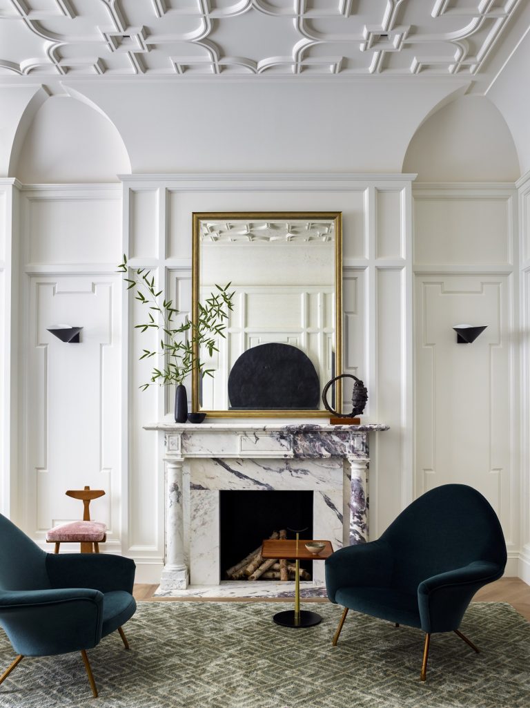Elegant living room in a West Village Townhouse with white paneled walls, a marble fireplace, and a large mirror above the mantel. Two dark teal armchairs face each other on a patterned rug, complemented by minimalist New York style decor.