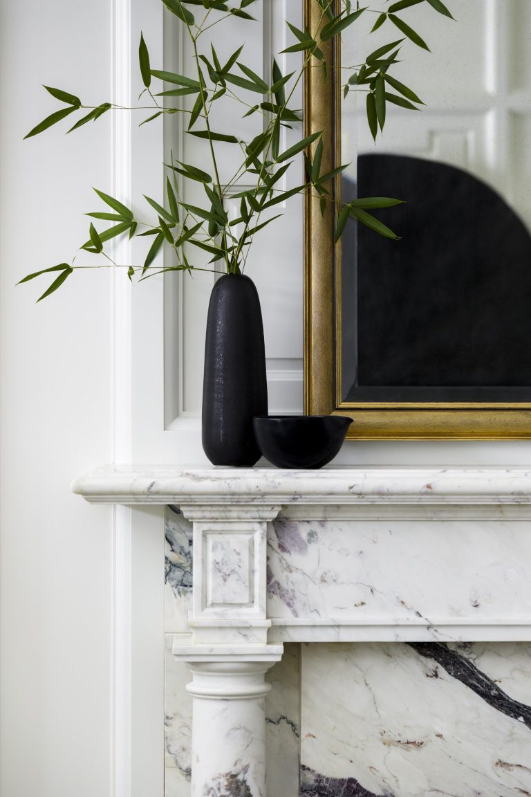 A black vase with green leafy branches and a small black bowl sit on a white marble mantel in this West Village Townhouse. Behind them, a large gold-framed mirror reflects the elegant, light-filled New York space—minimalist and stylish.