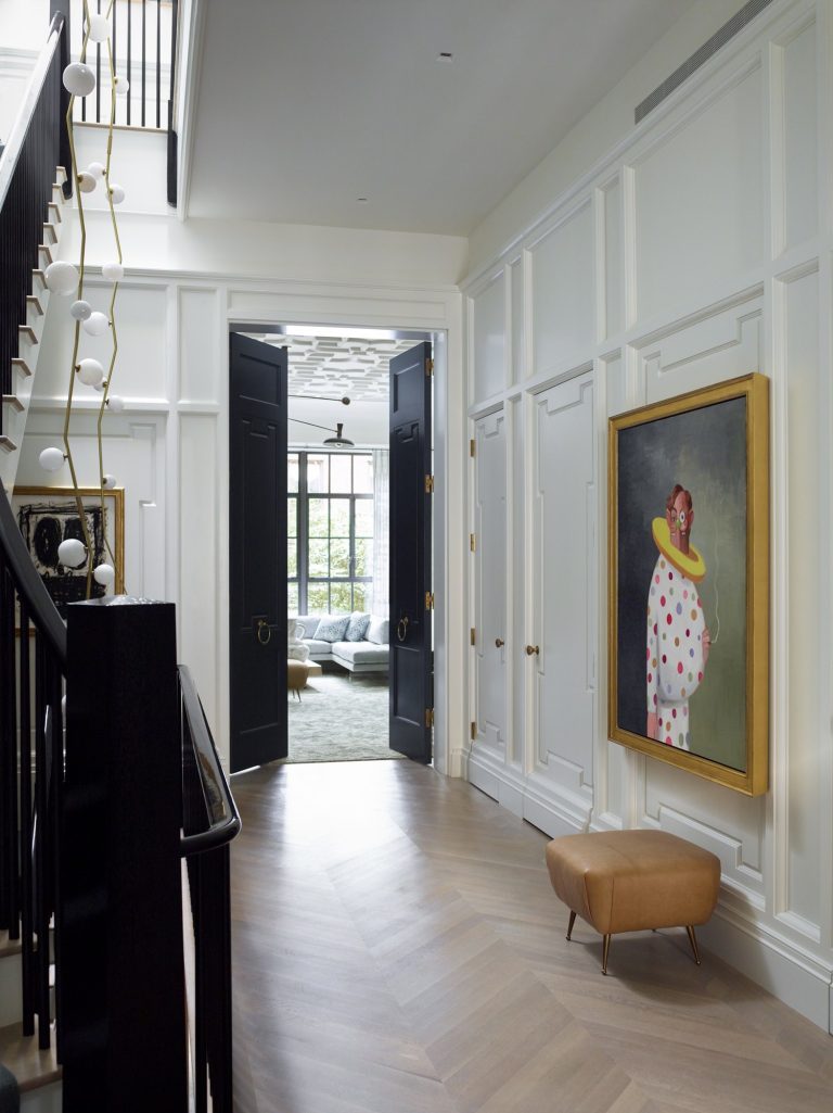 A modern West Village townhouse hallway with white paneled walls, herringbone wood floors, and black double doors opening to a bright room. A large framed painting hangs above a tan ottoman, while sculptural lighting lines the staircase.