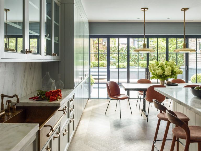 Bright, modern kitchen in a West Village townhouse with marble countertops, copper fixtures, and light gray cabinets. Pinkish chairs surround the table and island. Large windows overlook a green garden, while fresh flowers and vases decorate the space.