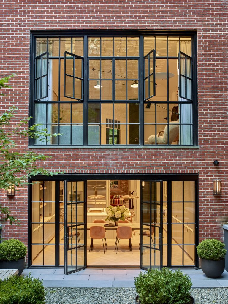 A modern West Village Townhouse with large black-framed glass doors on the first floor and matching windows above. The brightly lit interior features a dining table with pink chairs, a flower arrangement, and green potted plants framing the entrance.
