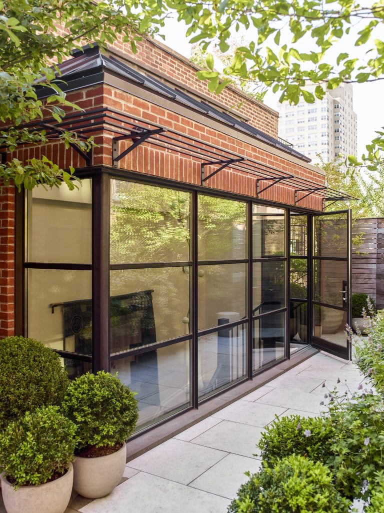 A modern townhouse extension with floor-to-ceiling glass windows and a black metal frame is attached to a brick exterior in New York’s West Village. Potted plants and trimmed bushes line the paved patio, framed by leafy trees and city buildings beyond.