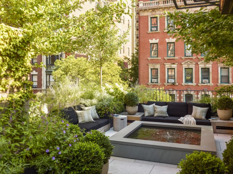 A lush urban patio in a West Village Townhouse features leafy plants, potted shrubs, and trees around a modern square fountain. Black cushioned sectionals offer cozy seating as New York’s red brick buildings rise in the background under a sunny sky.