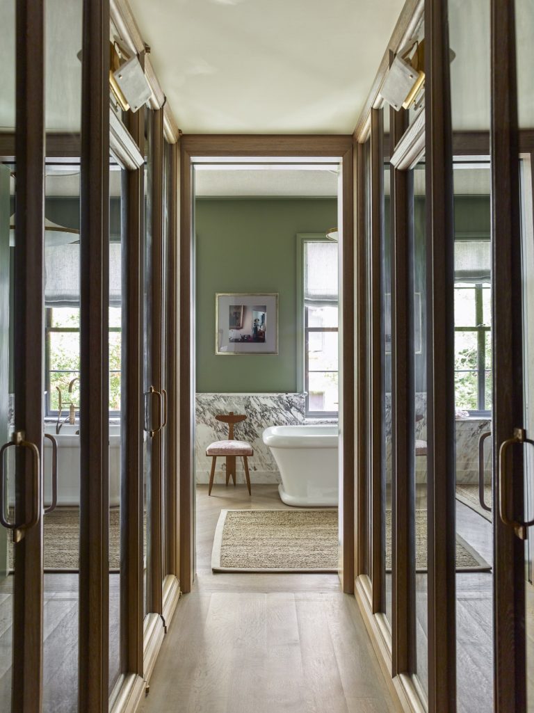A hallway with mirrored closet doors leads to a green-walled bathroom in this West Village townhouse. The bathroom features large windows, a freestanding white tub, marble wainscoting, and beige rugs on light wood floors bathed in natural light.
