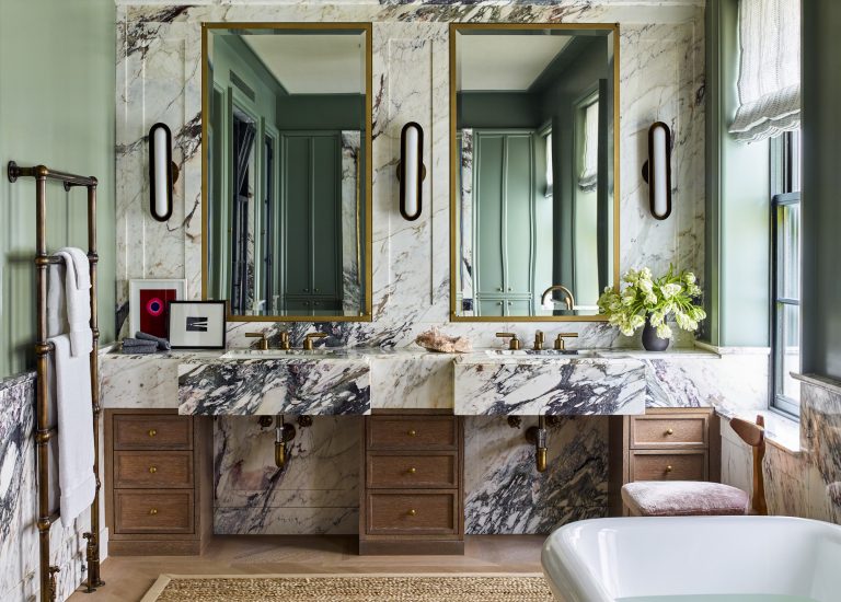 Elegant bathroom in a West Village Townhouse with two marble vanities, gold-framed mirrors, brass fixtures, and dark wood cabinets. Black sconces flank each mirror; a vase with greenery and sculpture grace the counter near a classic clawfoot tub.