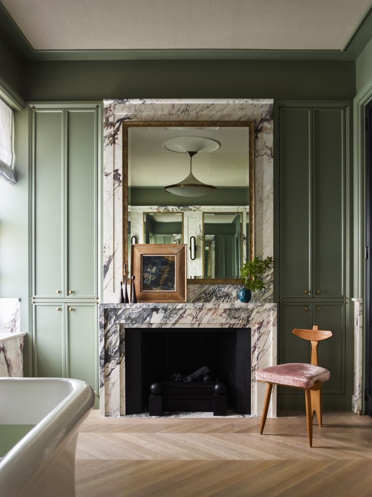 Elegant room in a West Village Townhouse, with sage green built-in cabinets flanking a marble fireplace. Above, a large gold-framed mirror reflects the space. The mantel features art and greenery; a wooden chair with a pink cushion sits to the right.