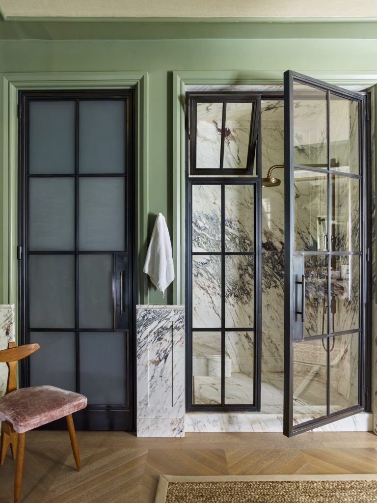 A modern bathroom in a West Village townhouse features sage green walls, black-framed glass doors, and marble surfaces. A towel hangs on the wall, while a velvet pink chair sits nearby—capturing chic New York style.