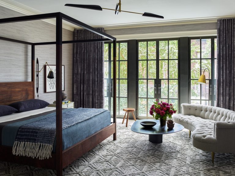 A modern bedroom in a West Village Townhouse features a dark wood four-poster bed, blue bedding, patterned rug, tufted cream sofa, round black coffee table with flowers, floor lamp, and large black-framed windows revealing green trees outside.