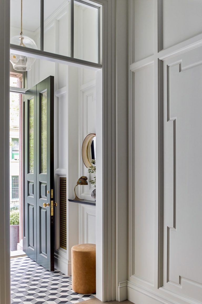 A stylish West Village Townhouse entryway features white paneled walls, a black door open to the outside, black-and-white geometric tile floor, a round mirror above a dark shelf with a vase, and a tan ottoman, all bathed in natural and pendant light.