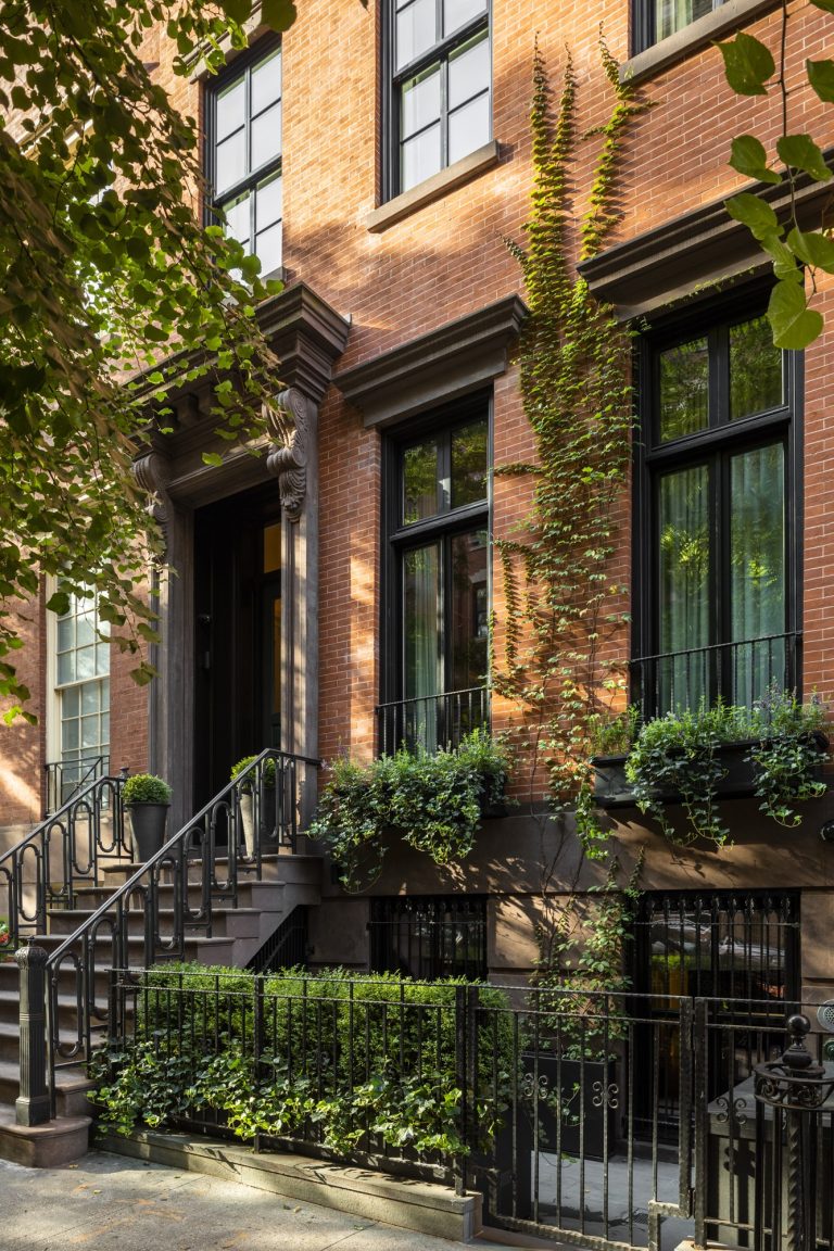 A classic red brick West Village townhouse with tall windows, black trim, and wrought iron railings. Ivy climbs the facade and window boxes overflow with green plants, creating a welcoming and elegant New York streetscape.