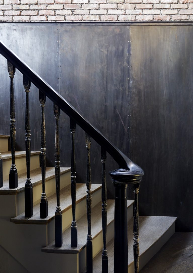 A close-up of a wooden staircase with black painted banister and spindles, some of which have chipped paint. The stairs have light wooden treads and risers. Behind the stairs is a wall with a dark, matte finish and a section of exposed brick above it.