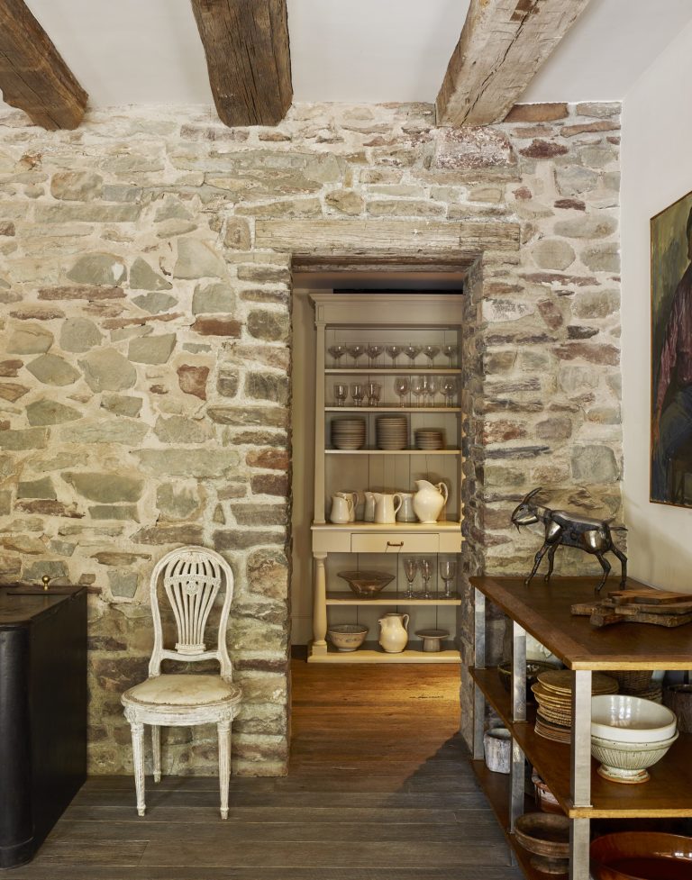 A rustic stone wall with wooden ceiling beams frames a doorway leading to a pantry with neatly arranged glassware, dishes, and pitchers. In the foreground, there’s a vintage white chair and a wooden table holding ceramics and a small horse sculpture.