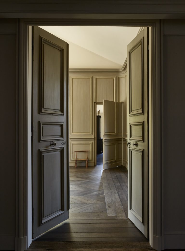 View through an open set of double wooden doors into a sunlit room with paneled beige walls, parquet wood flooring, and a small wooden stool in the center. Another doorway is visible in the background, with light streaming in.