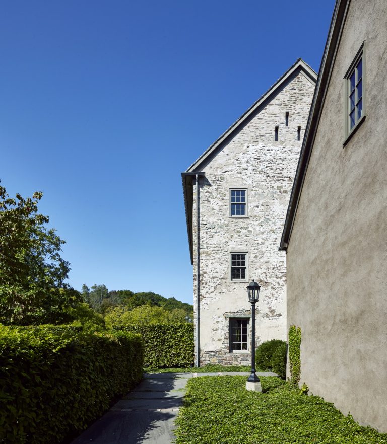 A stone and stucco building with three rectangular windows stands under a clear blue sky. A black lamppost sits by a path bordered by manicured green hedges. Lush trees and greenery are visible in the background.