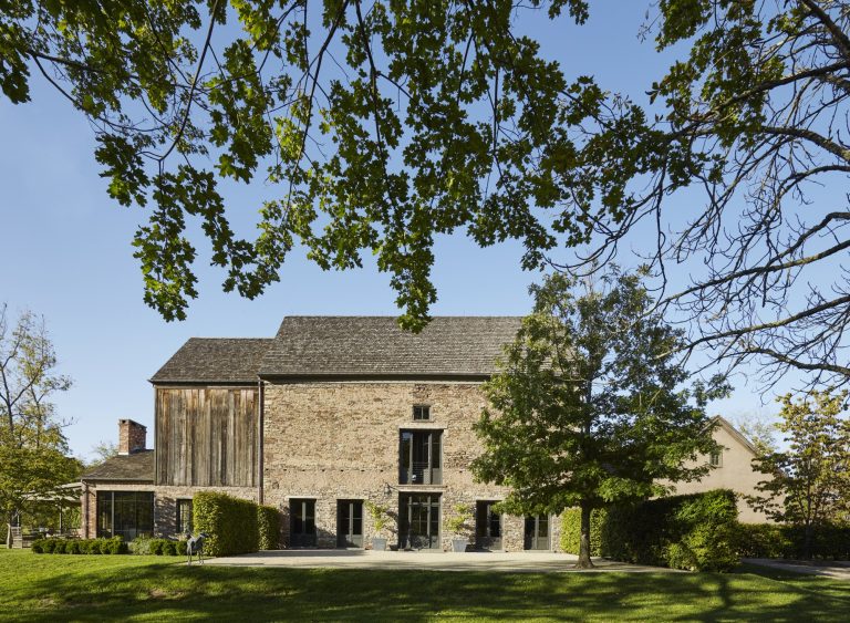 A large rustic house with stone and wood exterior stands under a clear blue sky. The foreground shows green grass and leafy tree branches framing the scene. The house features tall windows, hedges, and a few outdoor chairs on a paved area.