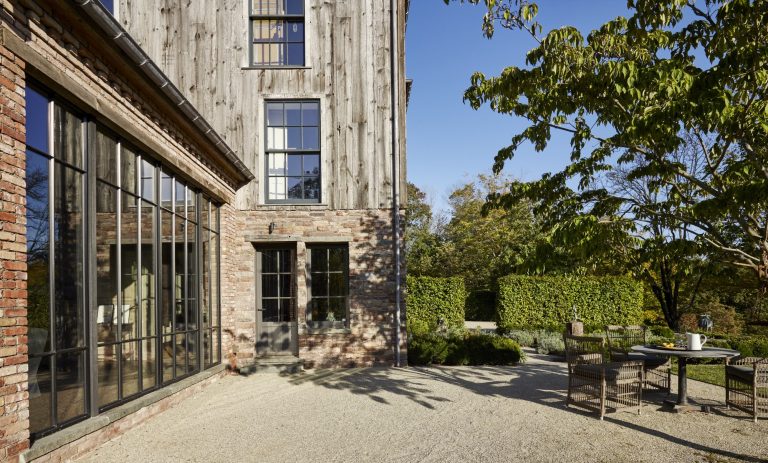 A rustic house with brick and weathered wood siding has large black-framed windows. A gravel patio with a table and chairs sits under leafy trees, surrounded by manicured hedges and greenery, with clear blue sky in the background.