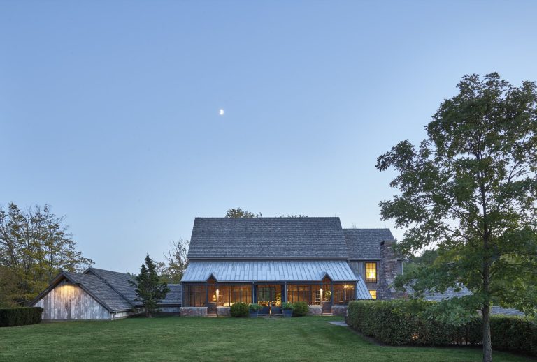 A large, rustic house with a steep gray roof and brick exterior sits on a green lawn at dusk. Warm lights shine from the windows. There is a detached garage on the left, tall trees around the property, and a clear sky with a small crescent moon above.