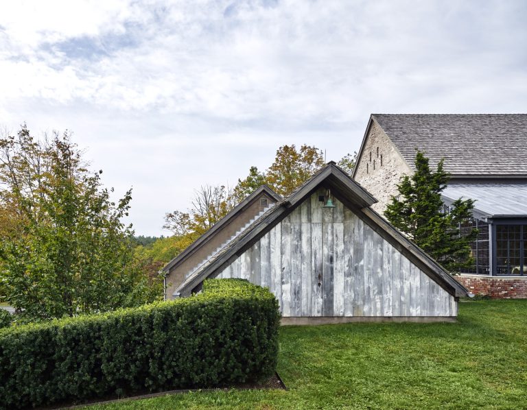 A small weathered wooden shed with a sloped roof stands on a green lawn, partially bordered by neatly trimmed bushes. Behind it is a larger stone building with a gray roof. Trees and cloudy sky are visible in the background.