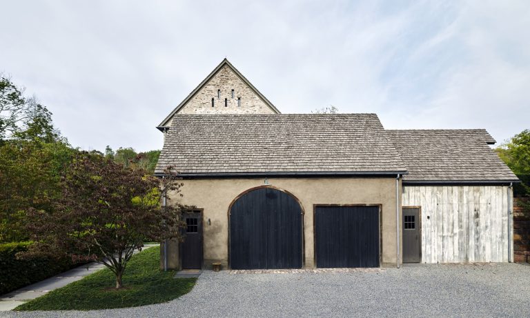 A rustic building with a sloped shingle roof, featuring a large black arched wooden door, a smaller black double door, and a white wooden panel section. Gravel covers the ground, and a tree stands to the left near a stone path. Green trees are in the background.