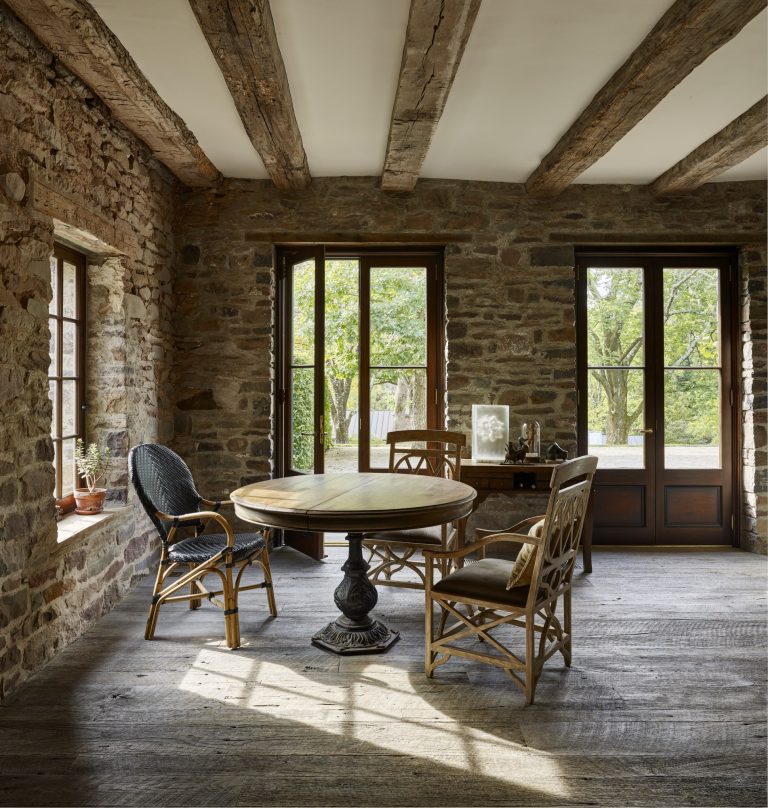 A rustic dining room with exposed stone walls, wooden ceiling beams, and large windows. A round wooden table with a black pedestal base is surrounded by four mismatched wooden chairs. Natural light fills the room, and greenery is visible outside.