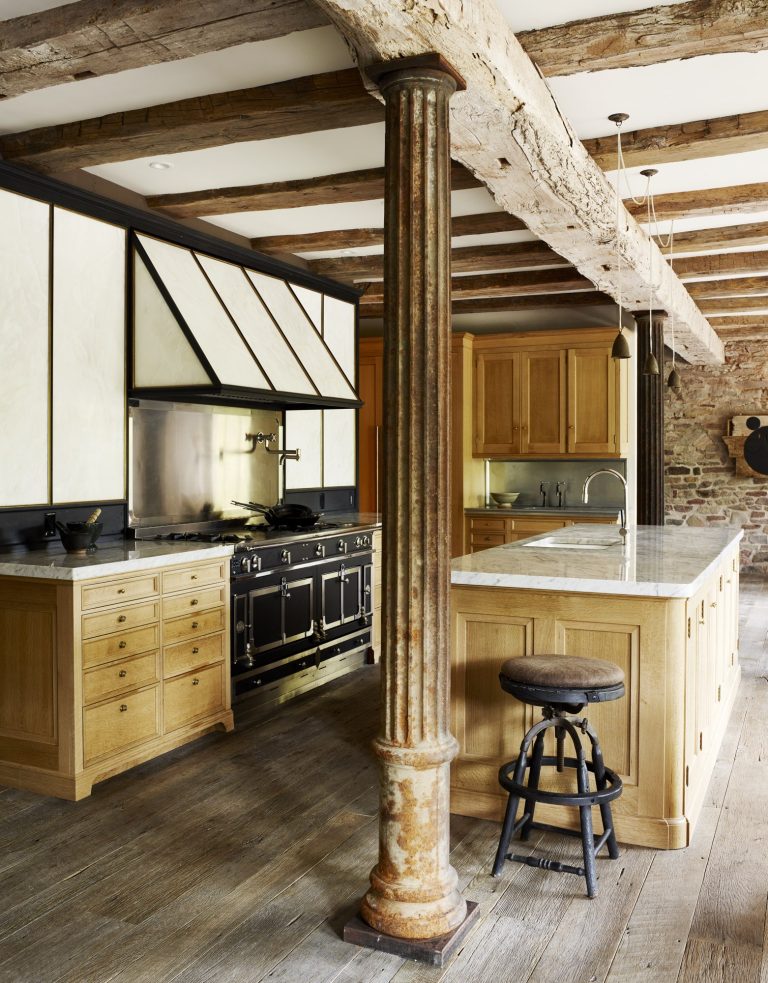 A rustic kitchen with exposed wooden beams, light wood cabinets, a marble-topped island, a black vintage stove, and a weathered metal column in the foreground. The floor is wide wooden planks, and natural light brightens the space.