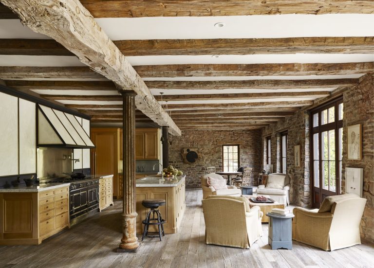 Rustic kitchen and living space with exposed wooden beams, stone walls, wide plank wood floor, and large windows. Light wood cabinets, a black range, a farmhouse island, wicker chairs, and cozy seating create a warm, inviting atmosphere.