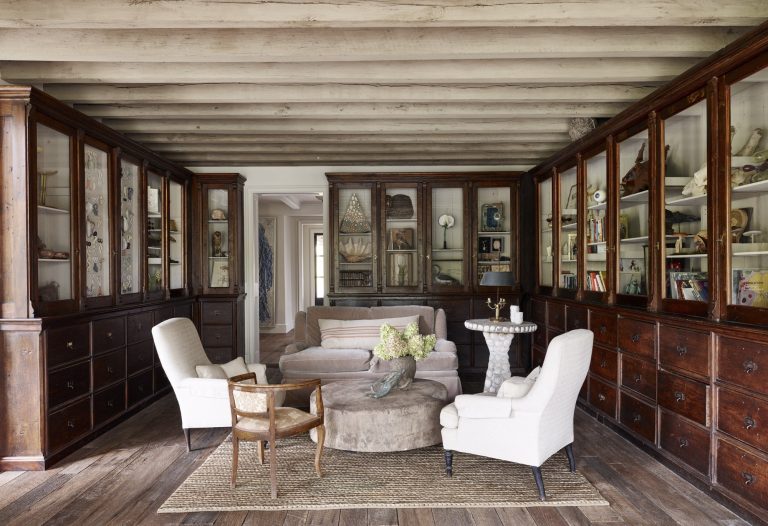 A cozy, rustic library with exposed wooden beams, glass-fronted cabinets lining the walls, a light brown sofa, three white armchairs, a round ottoman coffee table, a small wooden chair, and a woven rug on wood floors. Decor and books fill the cabinets.