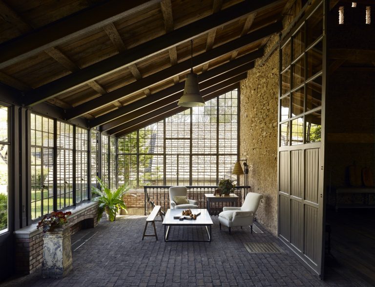 A sunlit rustic patio with a slanted wooden roof, exposed brick floor, and large metal-framed windows. There are two white cushioned chairs, a sofa, a coffee table with decor, potted plants, and a pendant light hanging overhead.