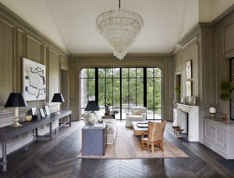 Elegant living room with herringbone wood floor, large glass doors overlooking greenery, neutral-toned furniture, a wicker chair on a rug, a white marble fireplace, abstract art, tall lamps, potted plants, and a dramatic crystal chandelier.