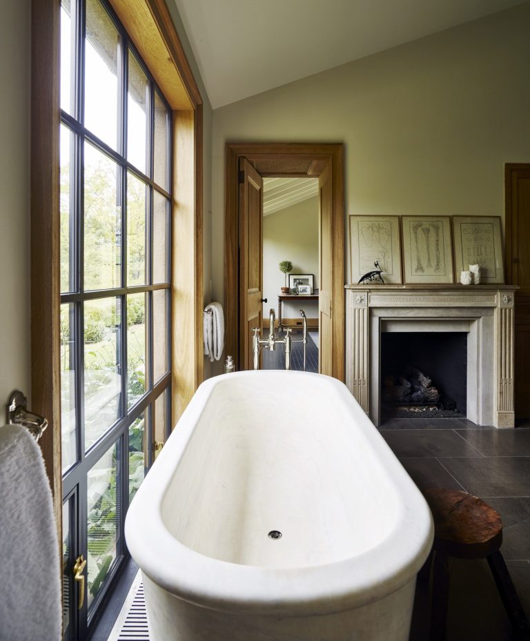 A white freestanding bathtub sits by tall windows with garden views. The room has a stone fireplace, art propped on the mantel, wood trim, taupe walls, and a side stool. Another room with a desk and chair is visible through an open wooden door.