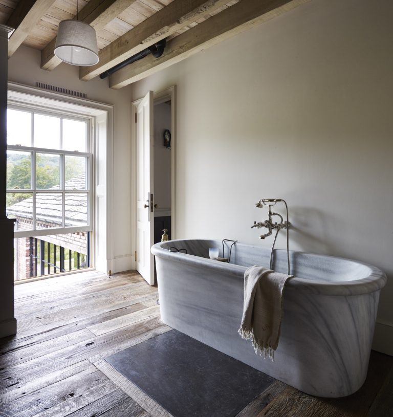 A rustic bathroom with a freestanding marble tub, a towel draped over the edge, wide plank wooden floors, a large window with a view of trees, exposed wood ceiling beams, and soft natural light filling the space.