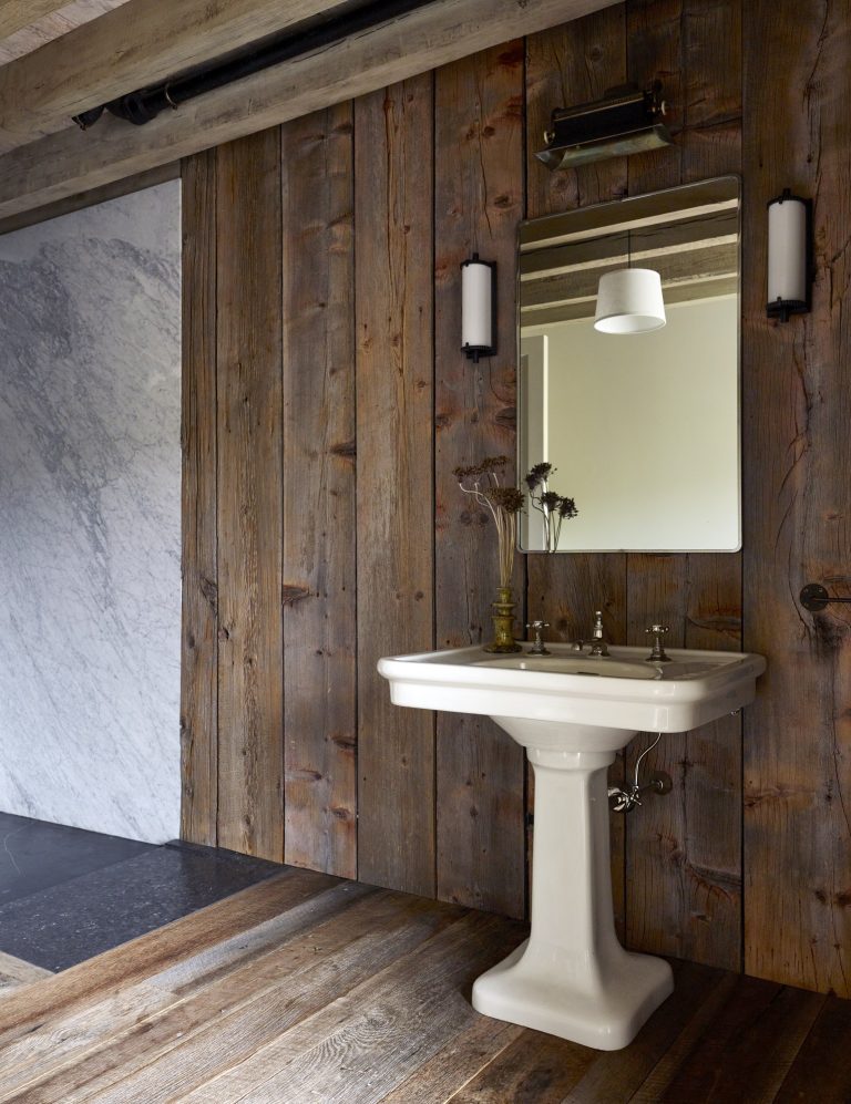 A pedestal sink with brass fixtures sits against a rustic wood plank wall. Above the sink is a rectangular mirror flanked by two wall sconces. A vase with dried flowers rests on the sink. The floor and ceiling are wood, and a marble wall is partially visible.