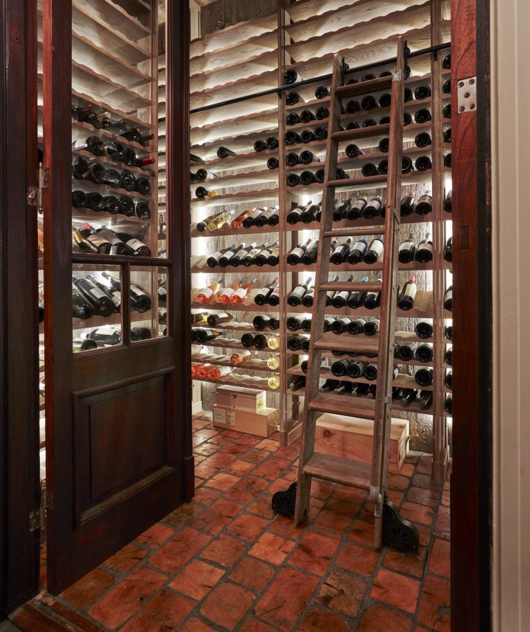 A wine cellar with brick flooring, wooden shelves filled with wine bottles, and a wooden door partially open. A wooden ladder leans against the shelves, and boxes of wine are stacked on the floor beneath the racks. Warm lighting illuminates the space.