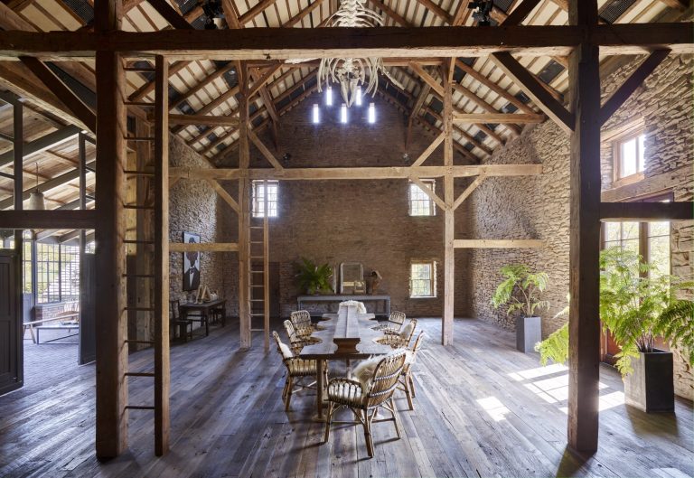 A spacious rustic dining room features exposed wooden beams, stone walls, and wide plank wooden floors. A round table with six cushioned chairs sits in the center. Large windows, potted ferns, and a modern chandelier create a bright, airy atmosphere.
