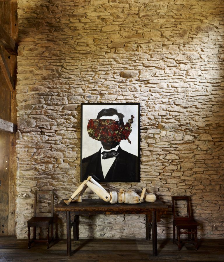A rustic stone wall displays a portrait of Abraham Lincoln with the US map obscuring his face. Below, a wooden table holds a mannequin with disjointed limbs. Two wooden chairs flank the table, creating a surreal, thought-provoking scene.