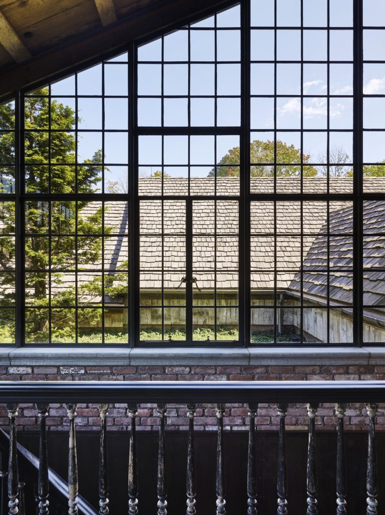 Large black-framed grid windows overlook a sloped wooden shingle roof and green trees. Sunlight streams in, illuminating a rustic brick wall and a dark wooden railing inside the building. The sky is bright blue with a few clouds.
