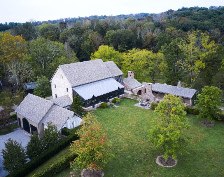 Aerial view of a large rustic house with a stone exterior, multiple sections, grey shingled roofs, and a wraparound porch. The property includes a detached garage, large grassy yard, surrounding trees, and is set against a wooded backdrop.