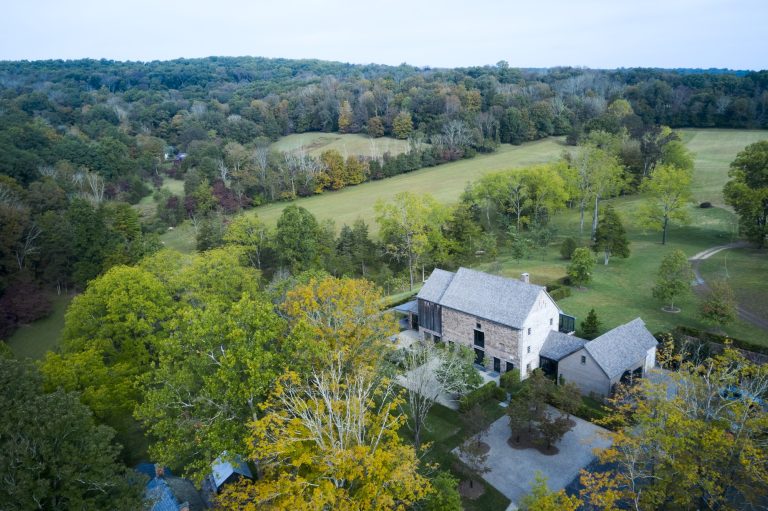 A large stone house with a gray roof stands among tall, leafy trees in early autumn. Surrounding the house are open grassy fields and dense woodlands, with rolling hills and a winding driveway visible in the background under a clear sky.