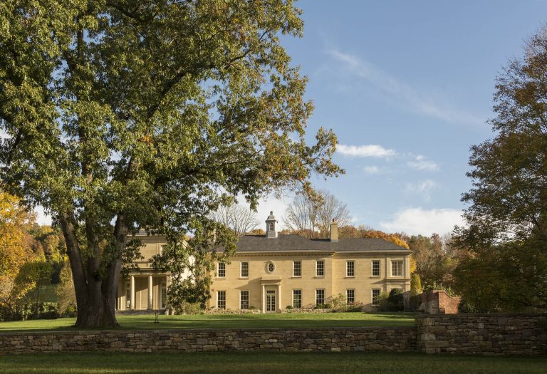 A large historic yellow house with a cupola, columns, and many windows sits behind a stone wall, surrounded by green lawns and tall trees under a blue sky with scattered clouds. Autumn foliage is visible in the background.
