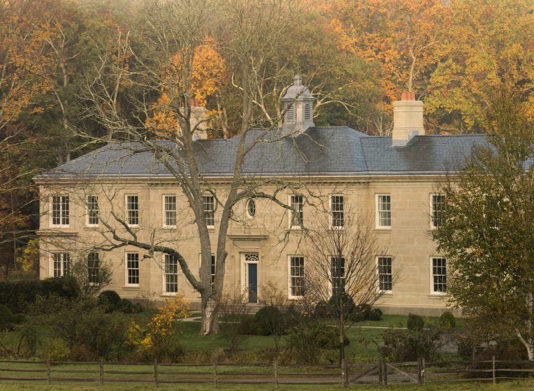 A large, beige colonial-style mansion with many windows and two chimneys stands amid autumn trees. A leafless tree is in front, and a wooden fence runs along the well-kept lawn. The background features colorful fall foliage.