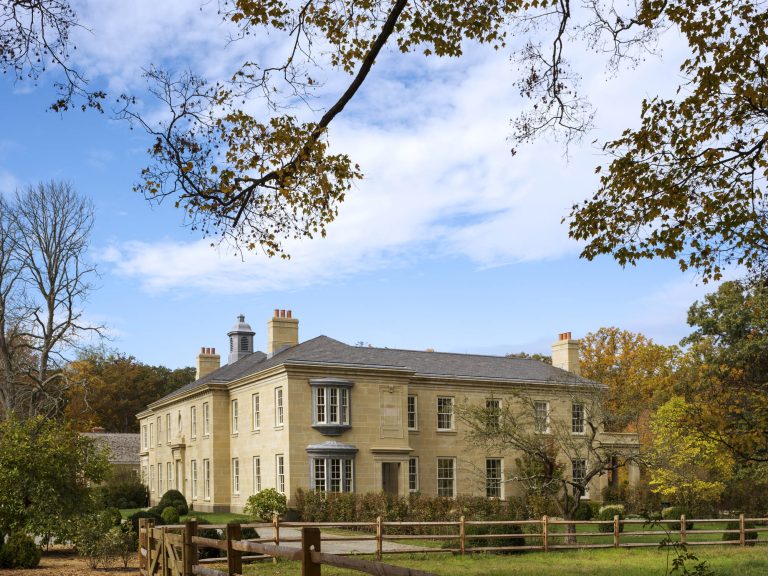 A large, elegant beige stone mansion with many windows and chimneys sits behind a wooden fence, surrounded by green grass, trees with autumn leaves, and a bright blue sky with scattered clouds.