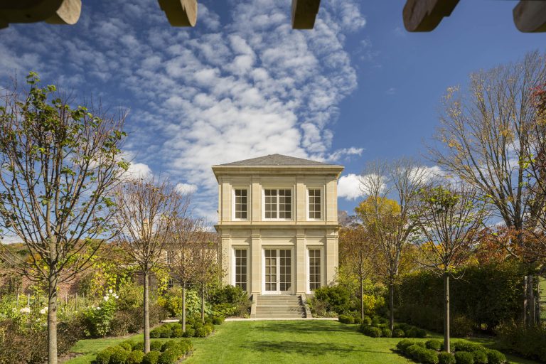 A symmetrical, two-story stone building with tall windows sits at the end of a manicured green lawn, framed by evenly spaced, pruned trees and hedges. The sky above is bright blue with scattered white clouds.
