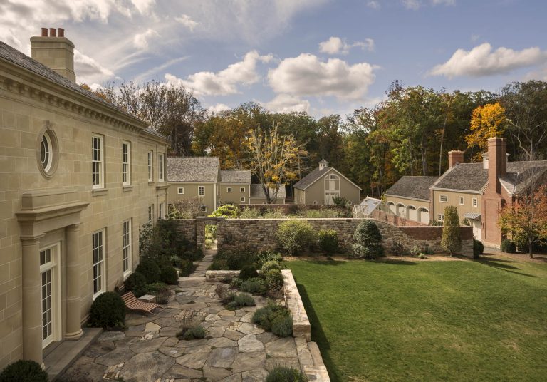 A large stone house with tall windows sits beside a manicured lawn and stone patio. Beyond a low stone wall, several outbuildings with gabled roofs are visible, surrounded by trees with autumn foliage under a partly cloudy sky.