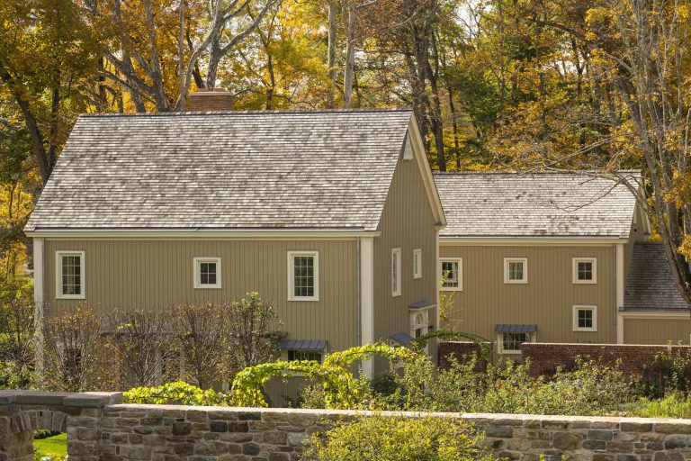 A beige, two-story house with white trim and a gray shingled roof stands amid autumn trees with golden leaves. The house has multiple rectangular windows and a chimney. In the foreground, a stone wall and green shrubs are visible.