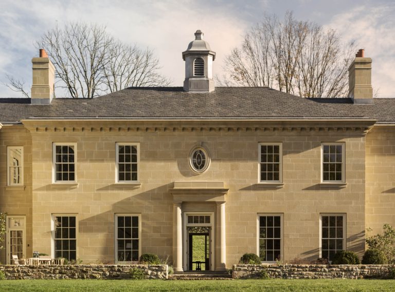 A large, symmetrical stone mansion features tall rectangular windows, a central entrance with columns and a pediment, an oval window above the door, two chimneys, and a small cupola on the slate roof. Leafless trees stand behind the house under a partly cloudy sky.