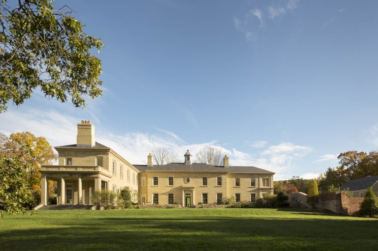A large, elegant yellow stone mansion with multiple chimneys and tall windows sits on a manicured green lawn. The house has a portico with columns on the left and is surrounded by autumn trees under a bright, clear blue sky.